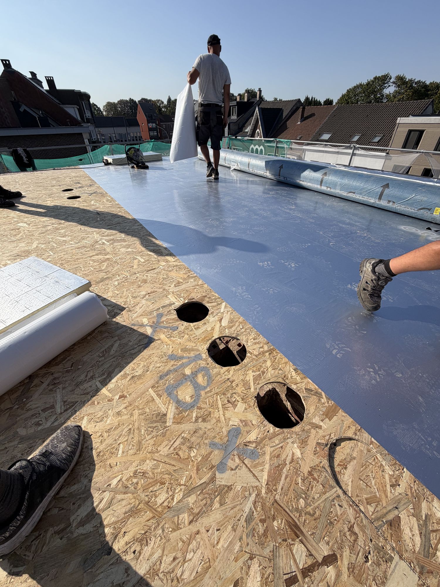 Het dampscherm wordt uitgerold op het OSB-dek van het plat dak in Gent, met de gaten voor de doorvoeren al uitgesneden