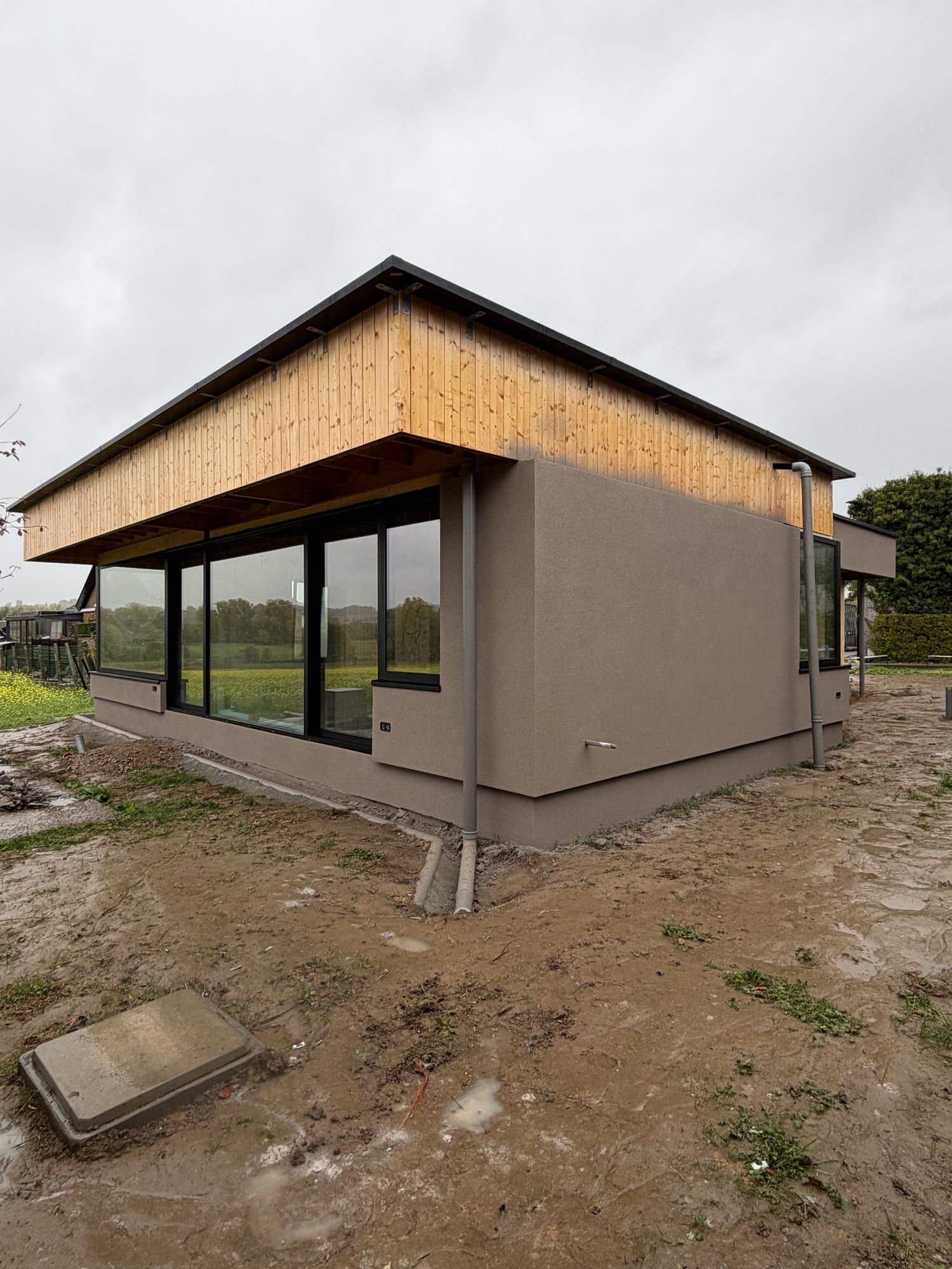 De afgewerkte gevelhoek in Brakel met taupe spuitkurk, het houten overstek erboven, een hoge glaspartij en de regenpijp