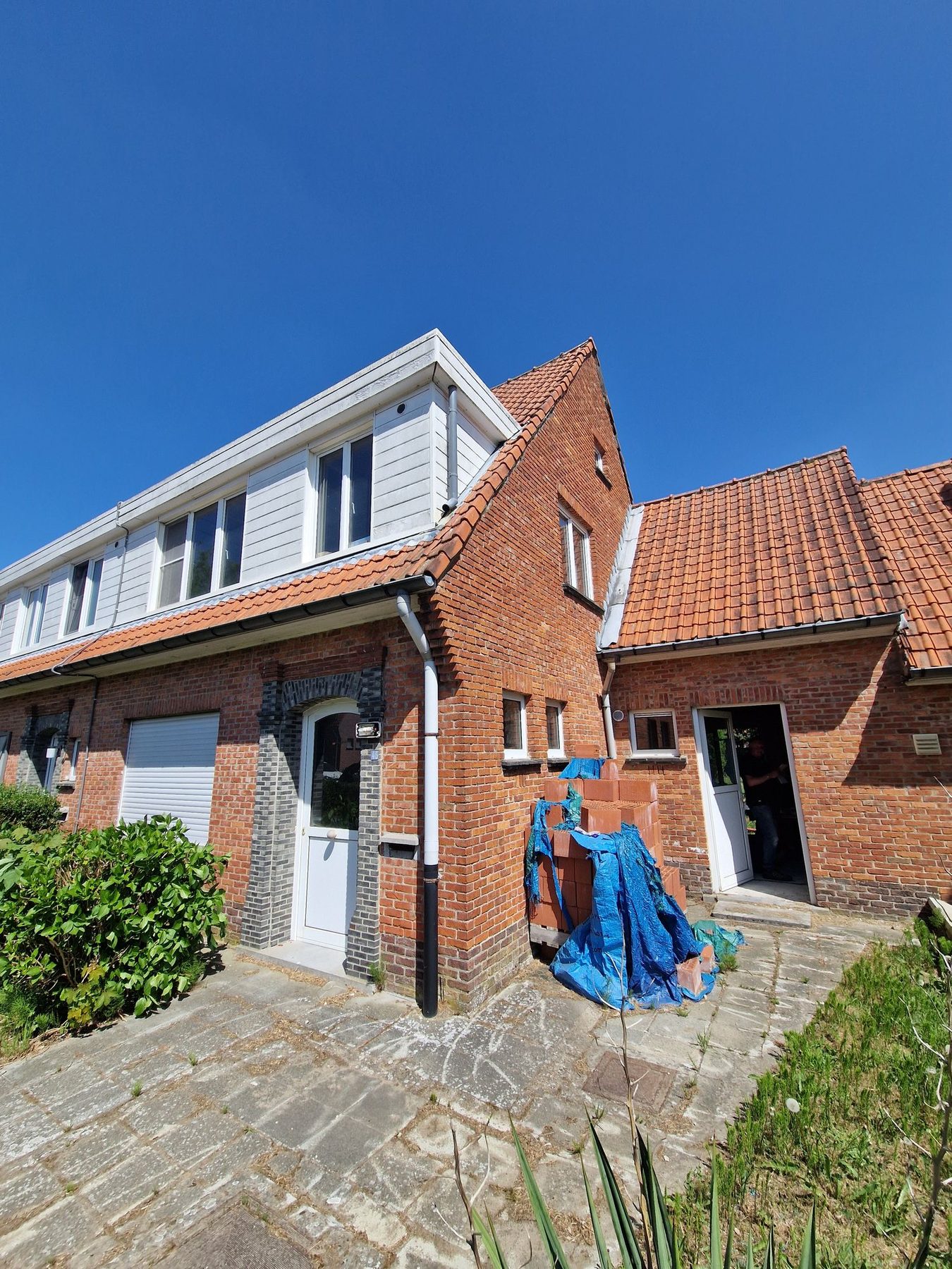 De rijwoning met aanbouw in Waasmunster voor de werken, met de oude dakbedekking en de onbehandelde baksteengevel